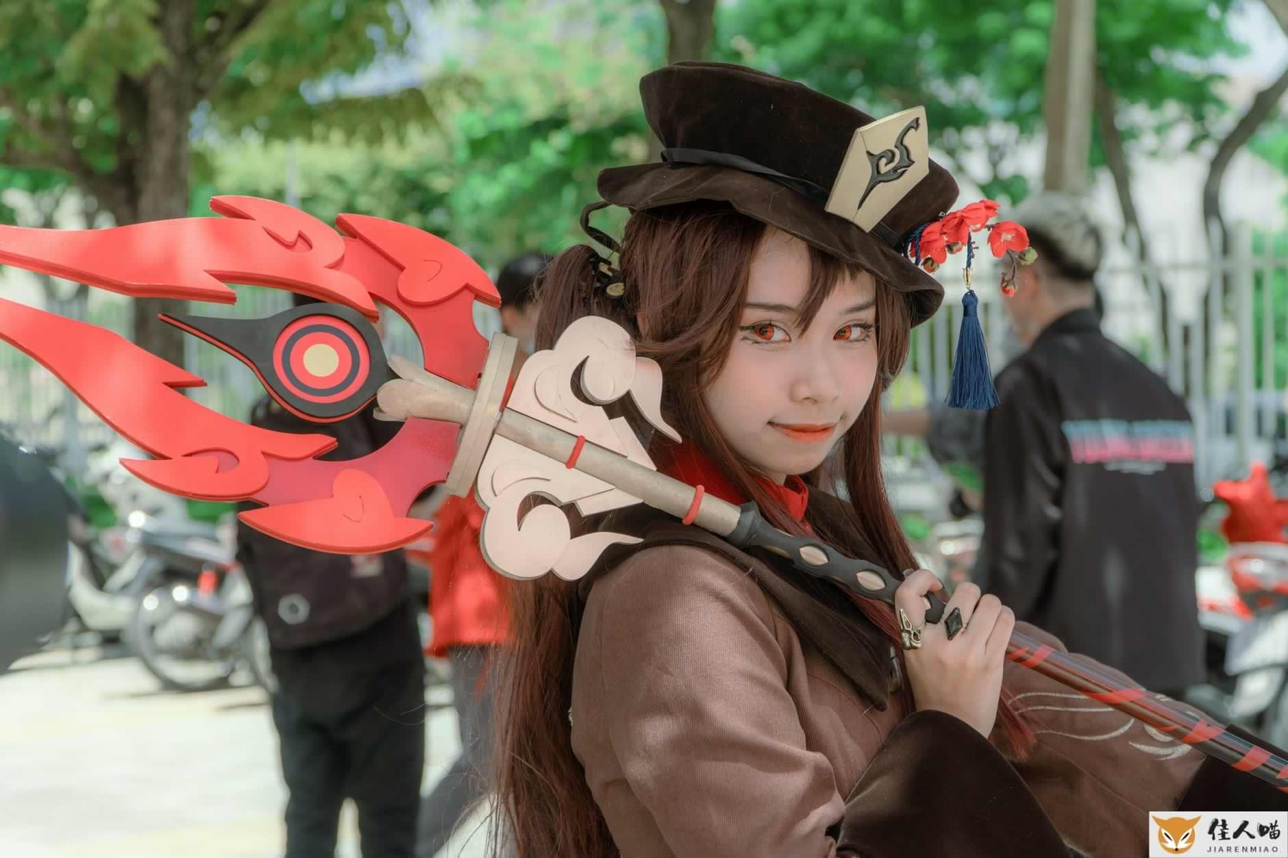 A female cosplayer dressed as Hu Tao from Genshin Impact poses outdoors with a detailed costume and weapon.